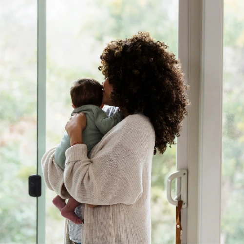 A mother with curly hair and a white sweater holds her newborn baby dressed in blue pajamas close as they stand in front of a window