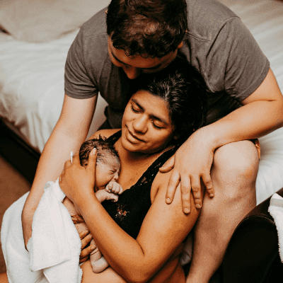 A father sits at the edge of a bed cuddling his wife and new baby after a home birth